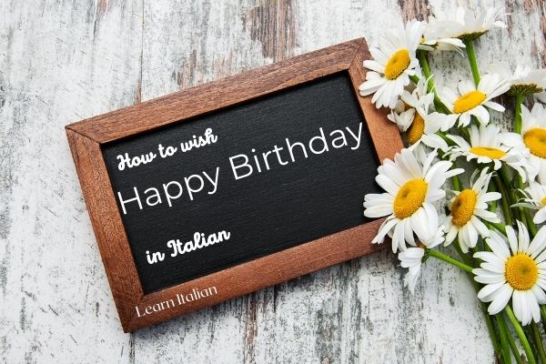 black board with how to say happy birthday in Italian wirtten on it and flowers beside
